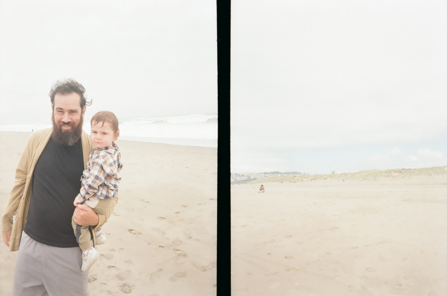 A bearded man wearing a beige jacket and gray shorts is holding a toddler in a plaid shirt while standing on a foggy beach with waves in the background.