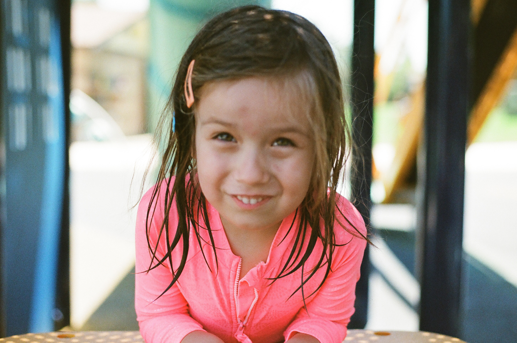 A young girl with wet hair and a pink top is smiling while leaning forward on a playground structure.