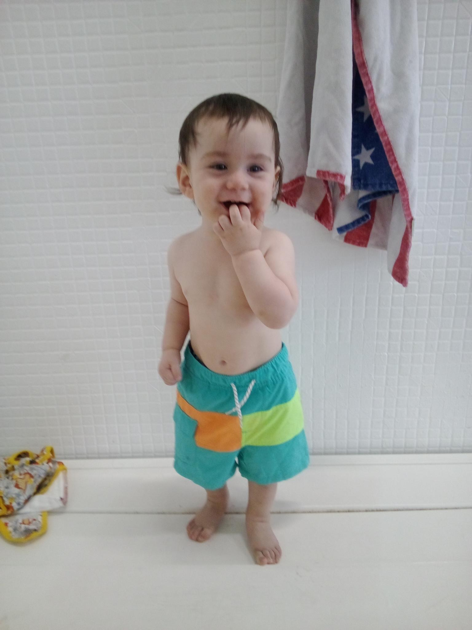 A toddler wearing colorful shorts stands against a tiled wall, smiling and holding a hand to their mouth.