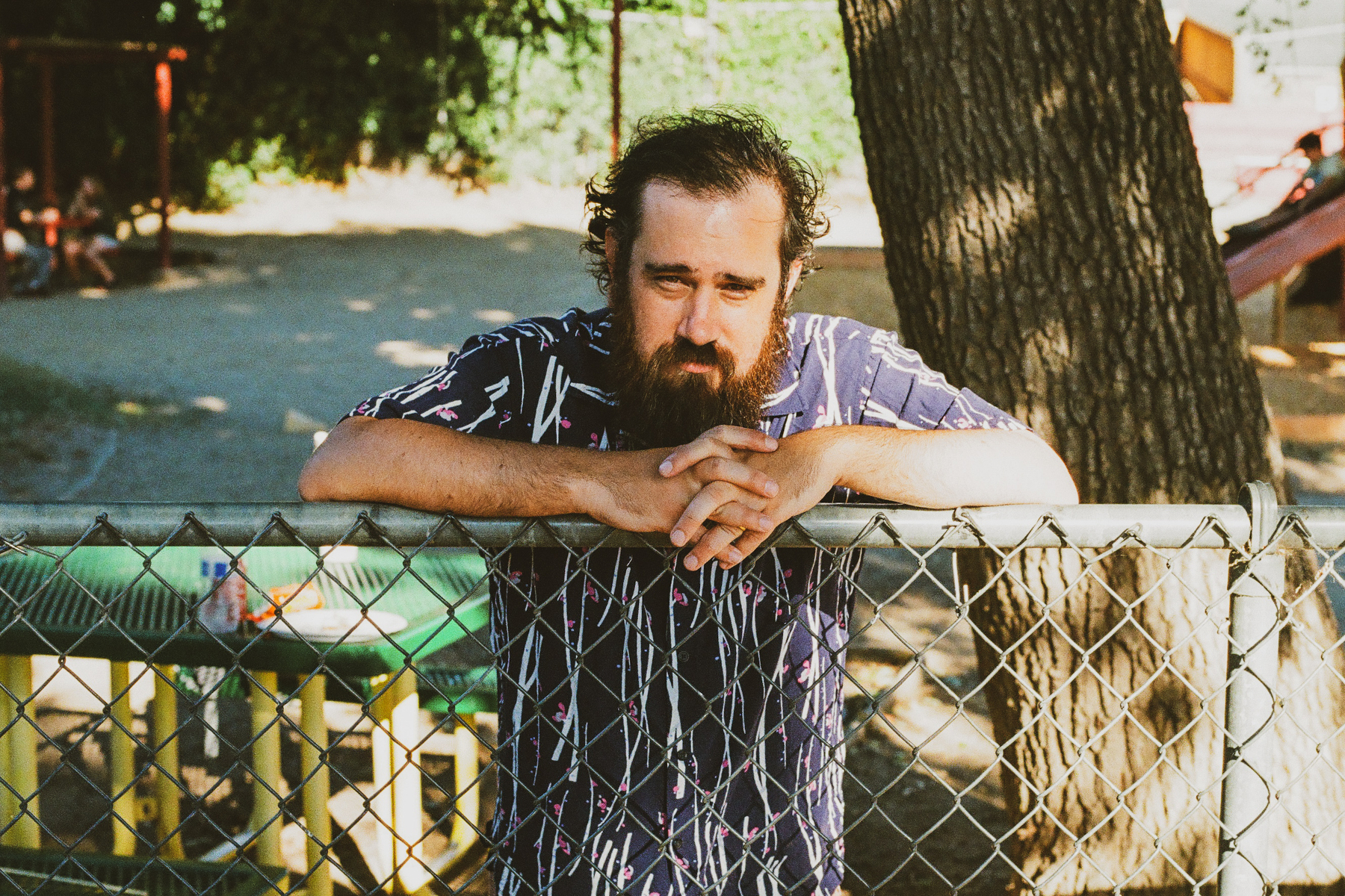 A man with a beard and patterned shirt is leaning on a chain-link fence outdoors, with trees and playground equipment in the background.