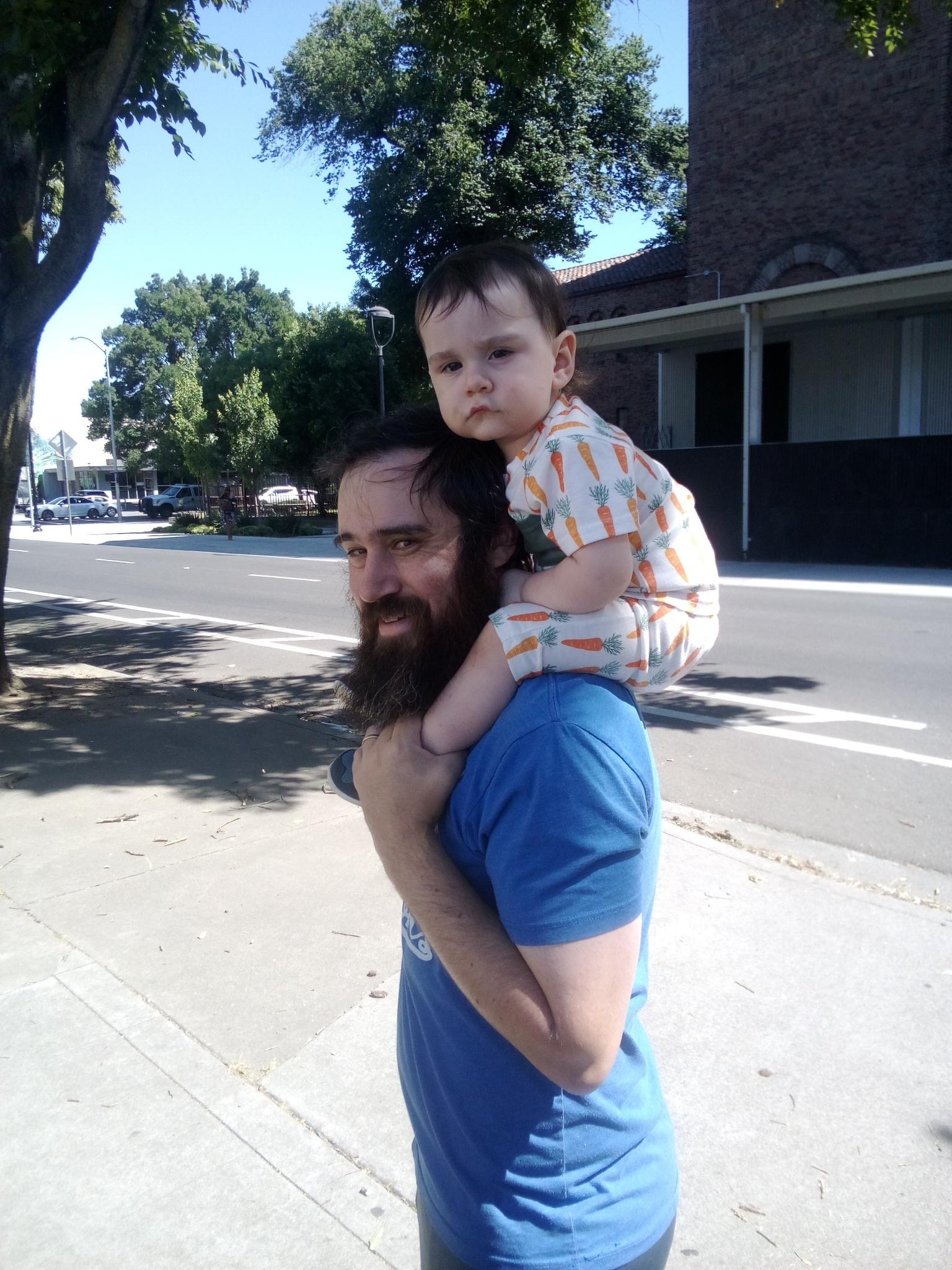 A man with a beard is carrying a baby on his shoulders while walking on a sidewalk.
