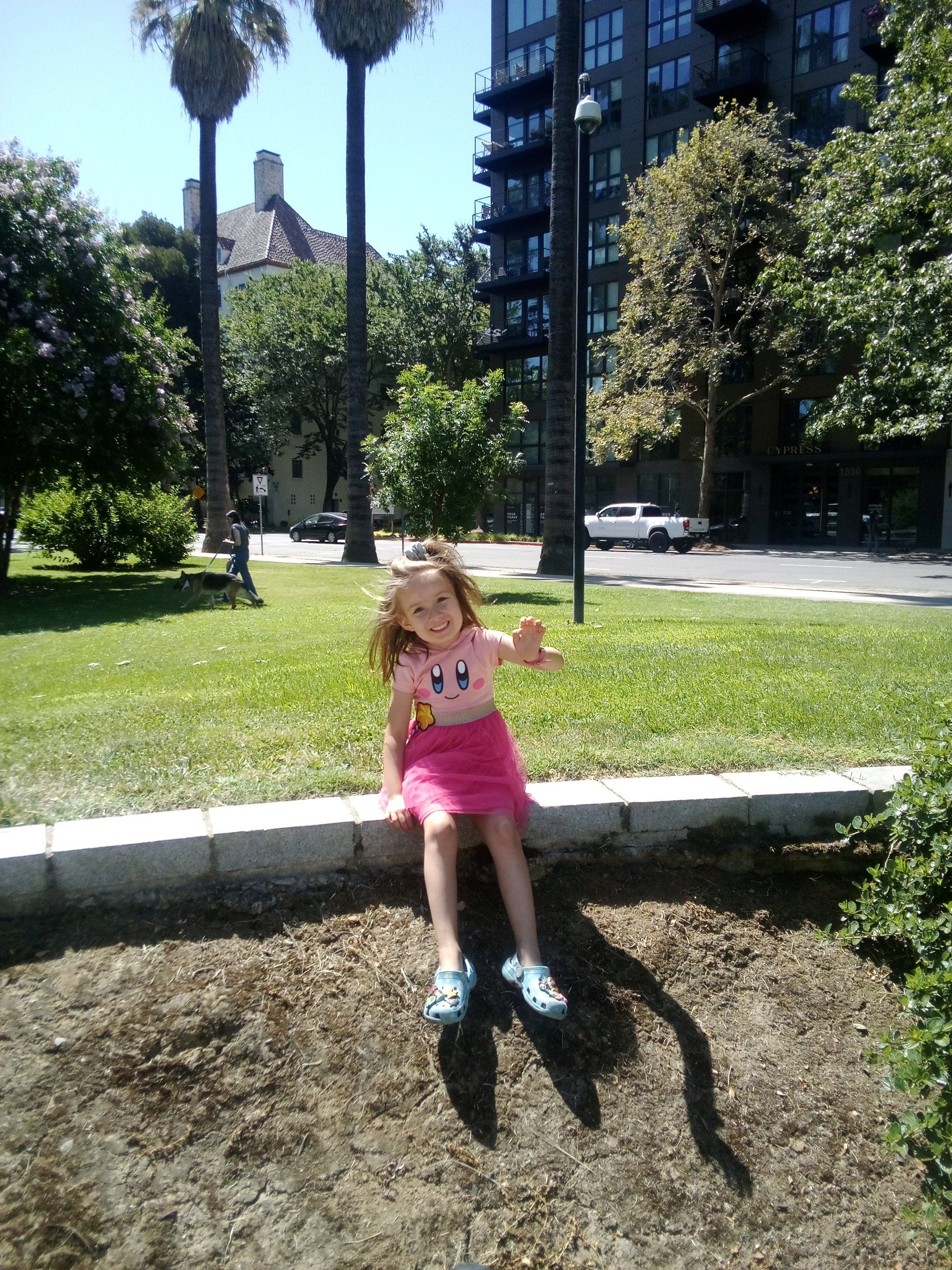 A young girl in a pink tutu dress and cartoon-themed shirt is sitting on a low stone wall in a sunny park, smiling and waving.