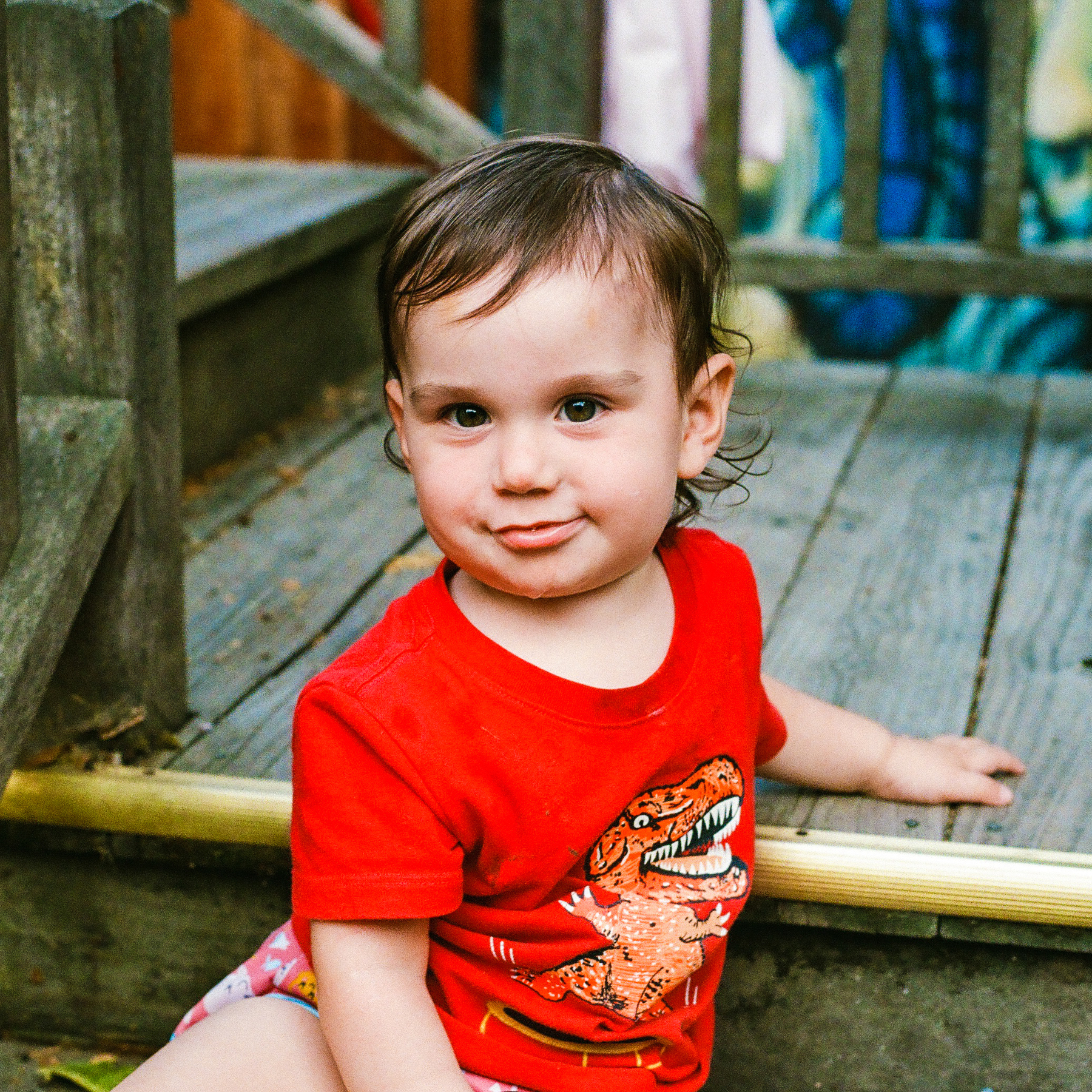 A toddler wearing a red dinosaur t-shirt is sitting on a wooden step outdoors, looking at the camera with a slight smile.