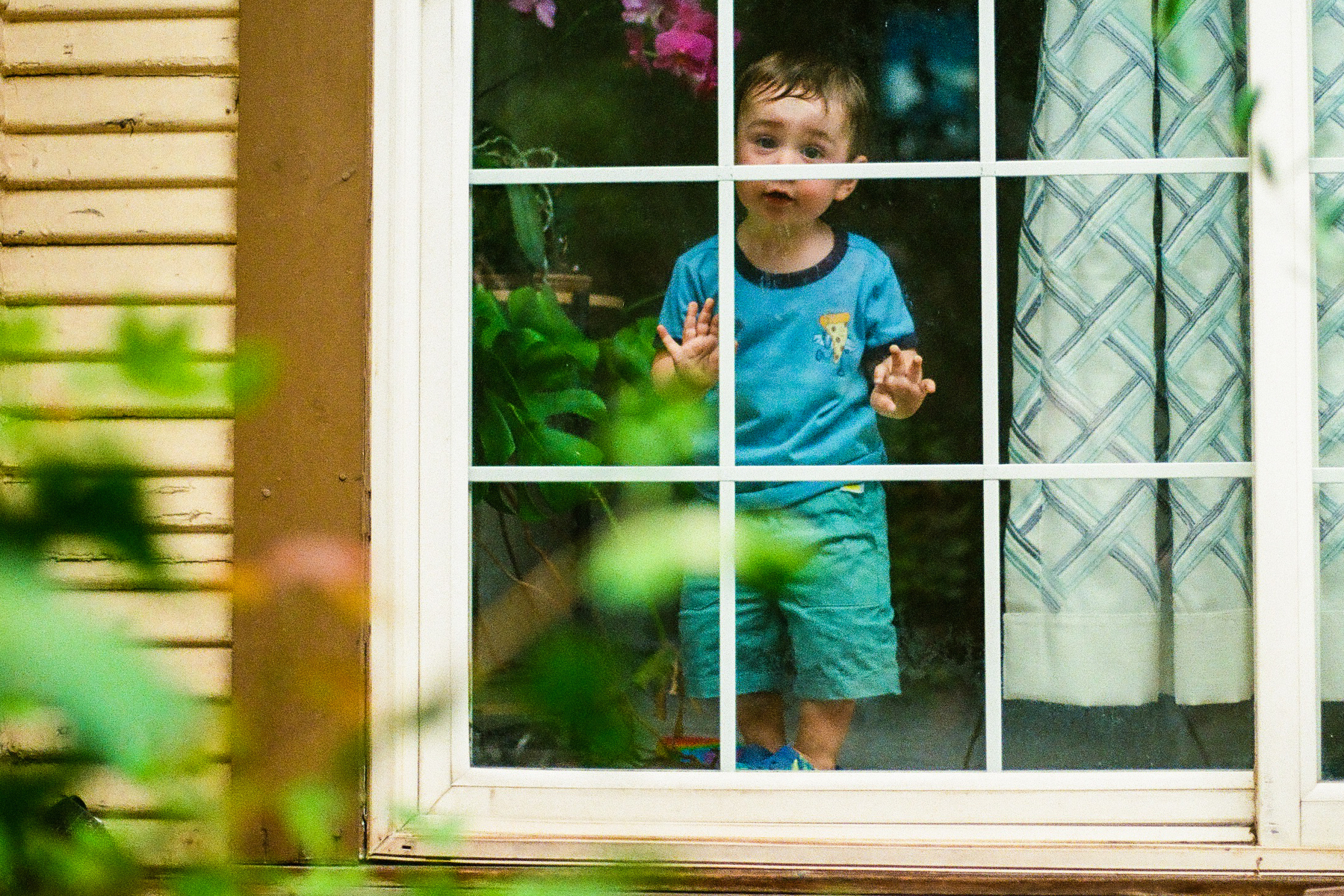 A young child wearing blue pajamas is standing behind a window, looking outside with a curious expression.