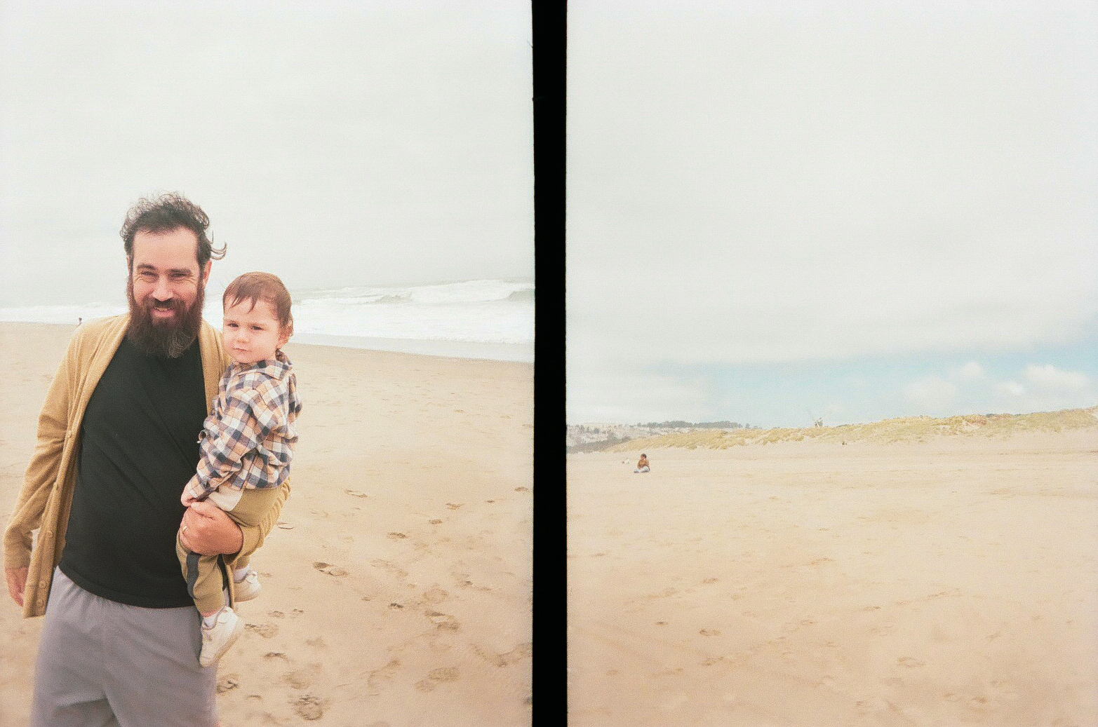 A man with a beard is holding a young child on a sandy beach with the ocean and cloudy sky in the background.