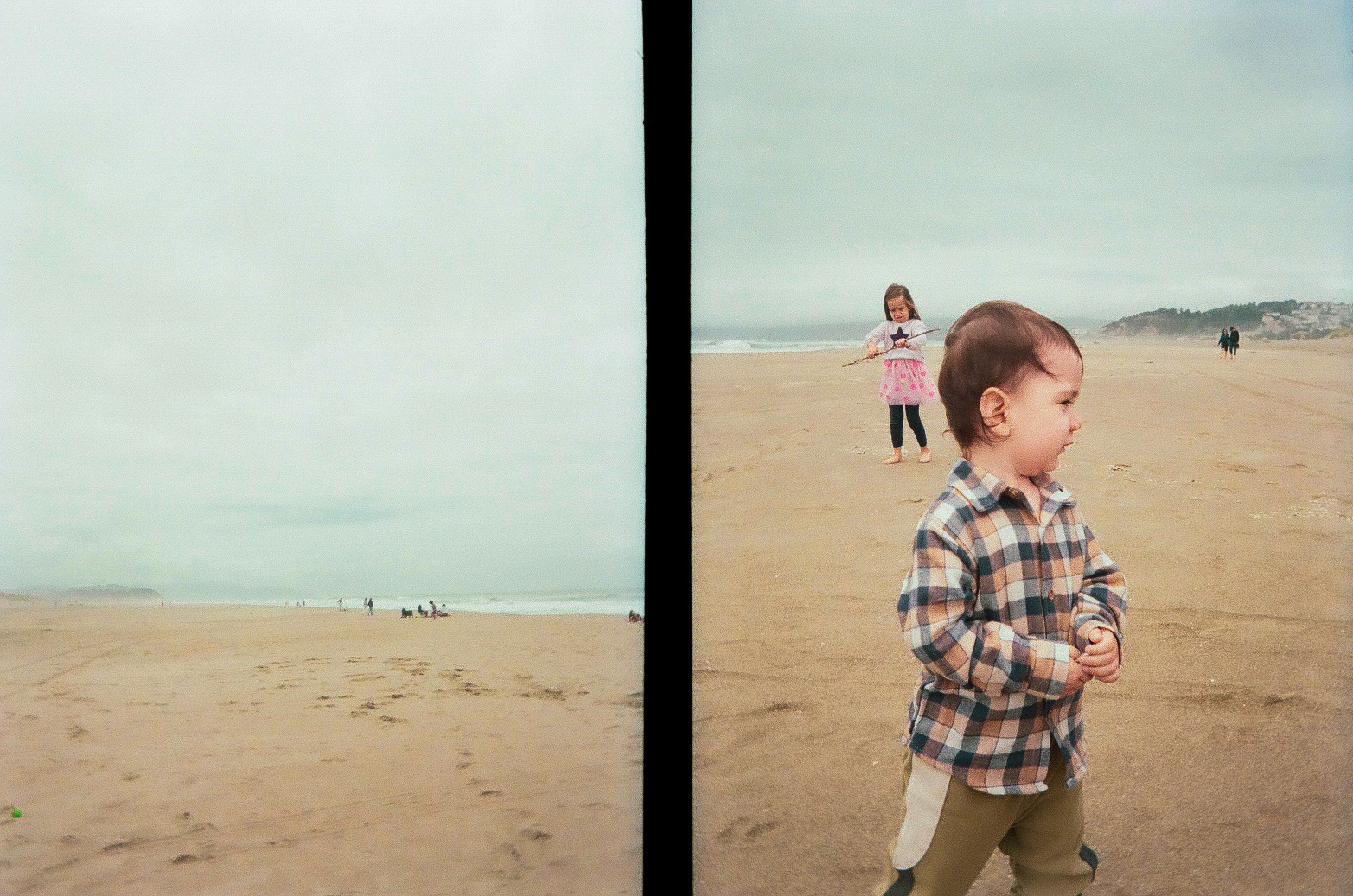 A toddler in a plaid shirt stands on a vast sandy beach with a girl and other distant figures in the background under a cloudy sky.
