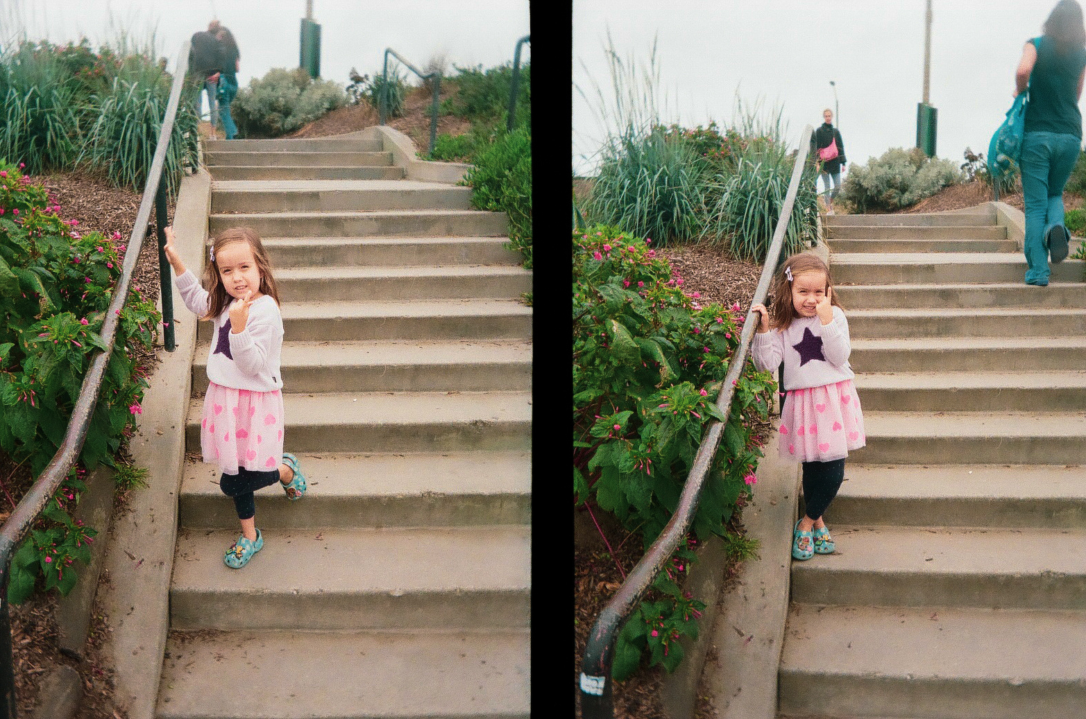 A young girl wearing a pink skirt and a purple star sweater is standing on outdoor concrete stairs, holding onto a railing and posing while people walk above her.