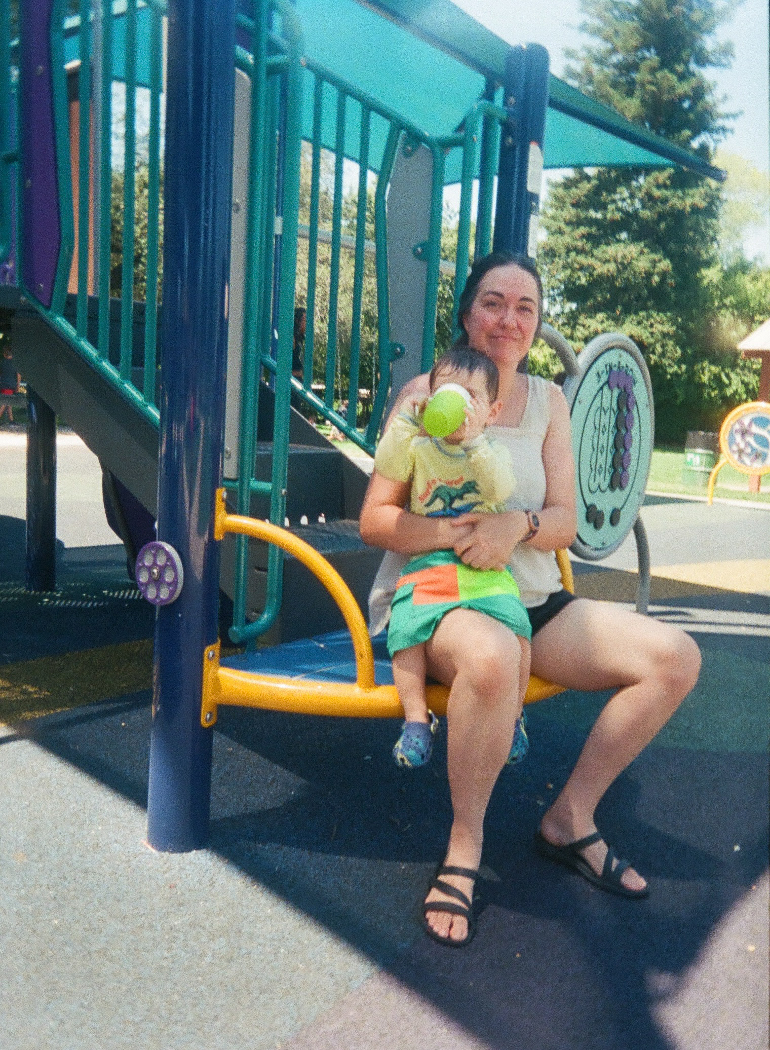 A woman is sitting on a playground swing holding a child who is drinking from a bottle.