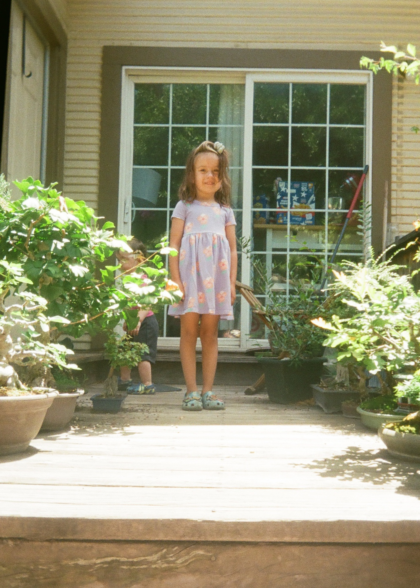 A young girl in a purple dress stands on a wooden deck surrounded by potted plants, with a sliding glass door and another child partially visible behind the plants.