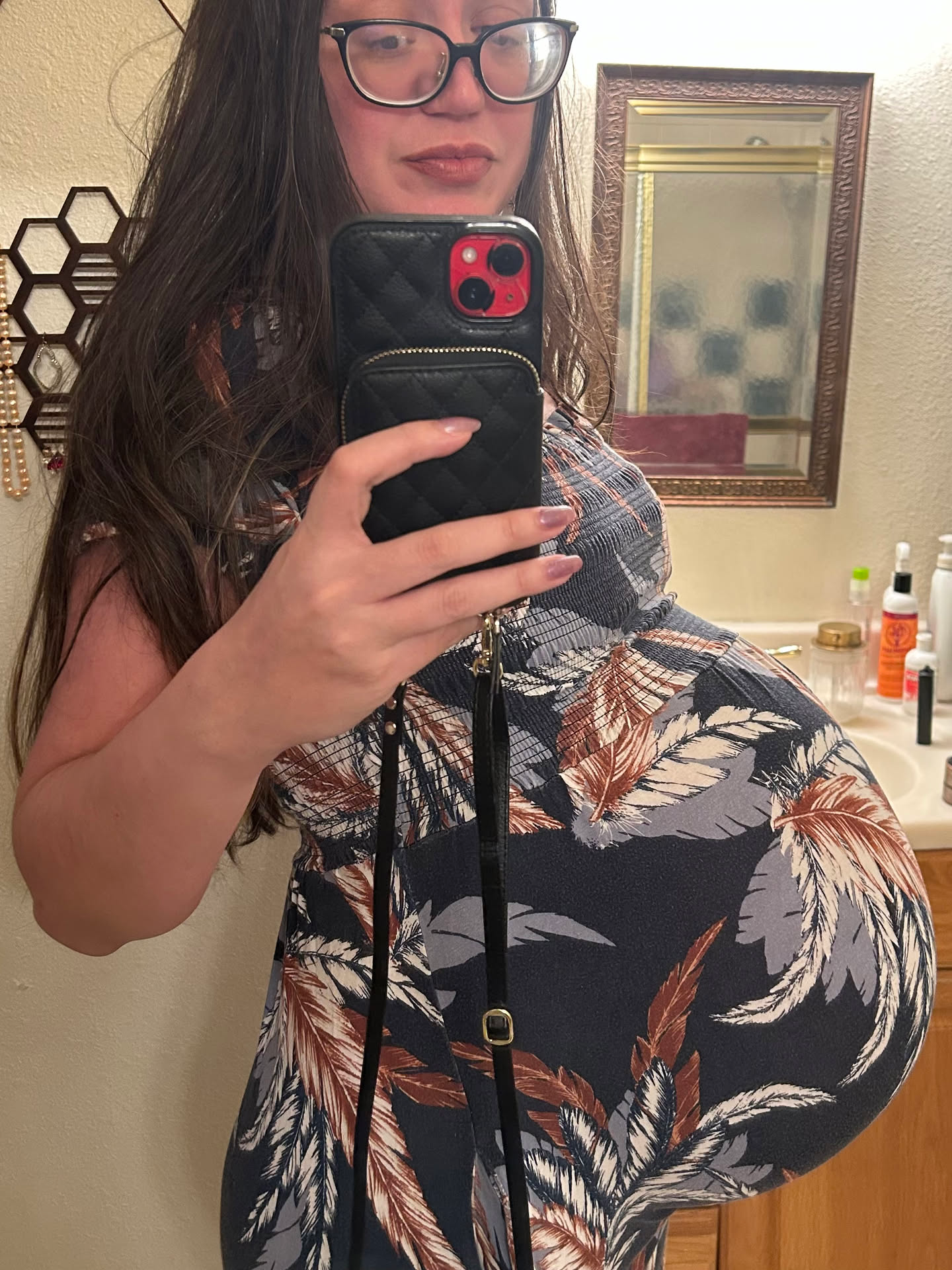 A woman with long dark hair and glasses is taking a mirror selfie in a bathroom while wearing a patterned dress that accentuates her large pregnant belly.